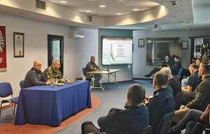 Policjanci siedzą na auli. Przy stole siedzi policjant i strażnik graniczny. Na tablicy wyświetlana jest prezentacja.