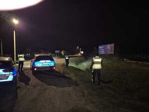 Troje policjantów  stoi przy dwóch radiowozach policyjnych na skrzyżowaniu drogi.