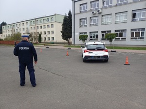 Obok policjanta na placu manewrowym samochód omijający ustawione pachołki.