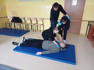 Dwie osoby udzielają pierwszej pomocy dwóm poszkodowanym, z których jedna leży na ziemi.