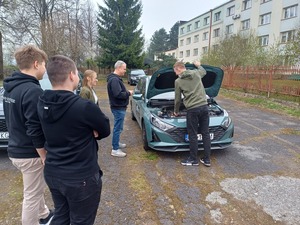 Kilka osób stoi przy samochodzie. Jedna  osoba trzyma podniesioną pokrywę silnika pojazdu.
