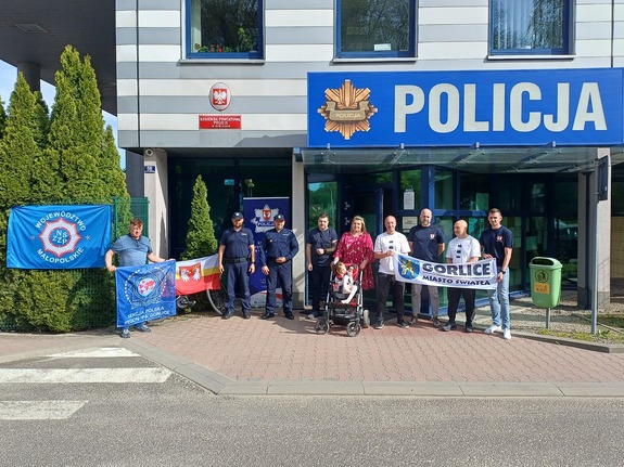 Przed komendą Policji w Gorlicach stoją uczestnicy rajdu charytatywnego wraz z dwoma policjantami oraz dzieckiem na wózku i jego rodzicami.