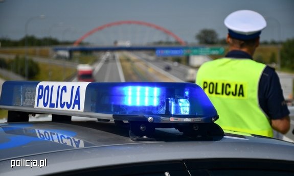 Policjant stojący tyłem obok radiowozu obserwuje drogę przed sobą.