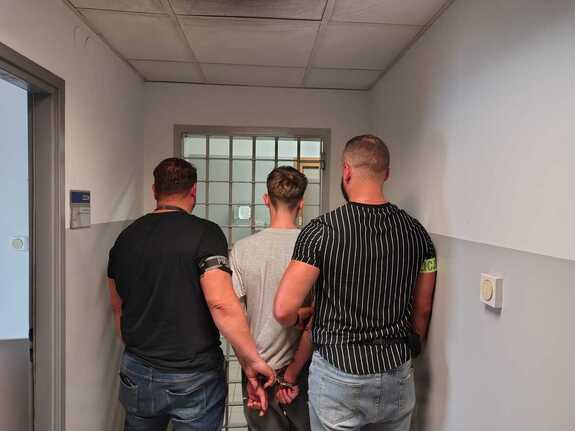 Mężczyzna stojący między dwoma policjantami stojącymi tyłem.