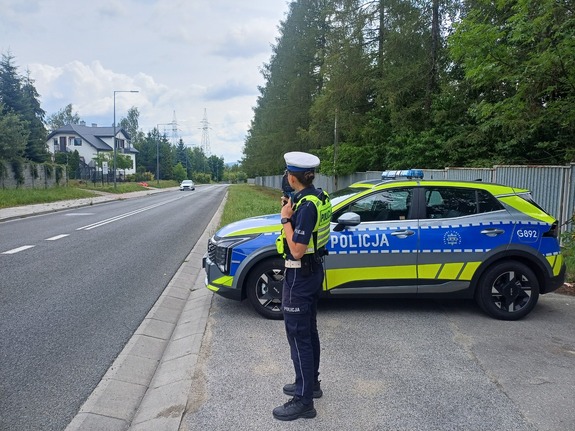Policjantka stoi obok radiowozu na poboczu drogi i mierzy prędkość jadącym  samochodom.