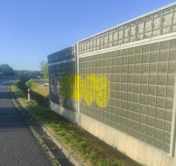 Przy drodze stoją ekrany dźwiękochłonne. Na powierzchni  ekranów naniesione malowidła w kolorze żółtym.