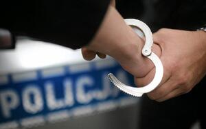 Na ręce zakładane są kajdanki. W tle napis Policja.