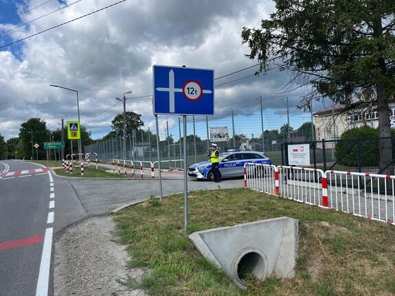 W pobliżu szkoły przy drodze stoi policjant przy radiowozie.