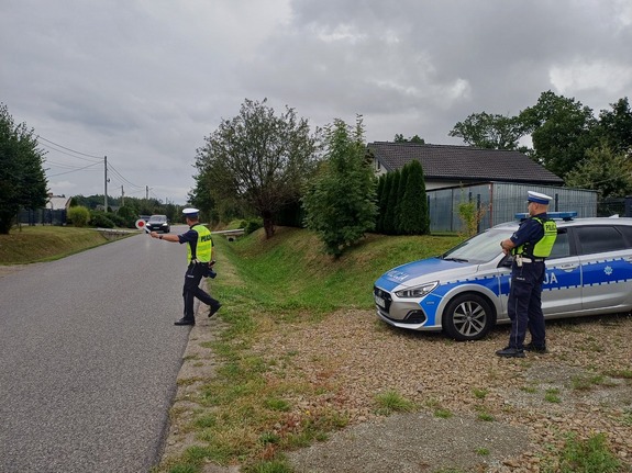 Dwóch policjantów przy radiowozie na poboczu drogi. Jeden policjant zatrzymuje jadący pojazd.