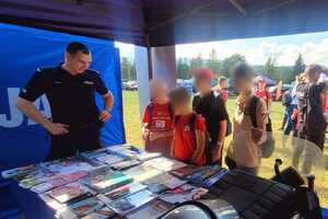 Policjant i dzieci na stoisku Policji. Na stole leżą ulotki profilaktyczne i sprzęt policyjny.