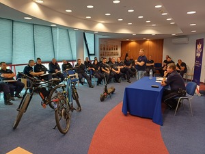 Policjanci siedzą na auli i słuchają szkolenia. Przy stole siedzi policjant, a przed stołem stoją dwa rowery i hulajnoga.