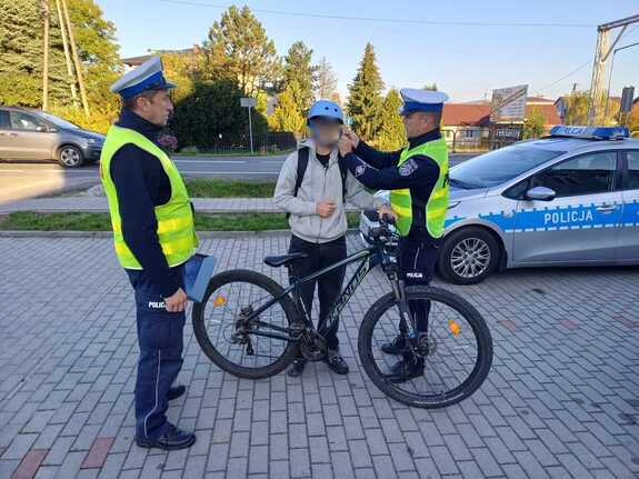 Policjant pomaga zapiąć kask chłopcu, który trzyma rower. Obok stoi drugi policjant, a w tle radiowóz.