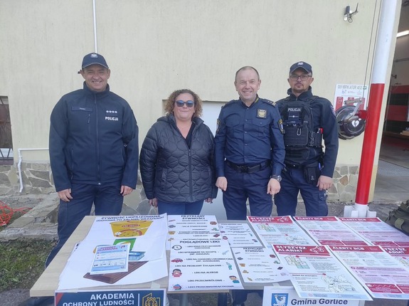 Przy stole z ulotkami informacyjnymi stoją policjanci i inne osoby.