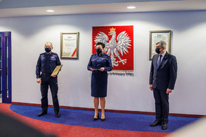 Komendant Powiatowy Policji wraz ze swoim Zastępcą i policjantem odchodzącym na emeryturę stoją w szeregu. W tle na ścianie Godło Polski.
