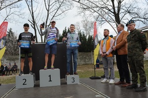 Na podium stoi trójka zwycięzców biegu trzyma w ręce puchary i  nagrody. Obok podium po prawej stronie stoją w rzędzie trzej mężczyźni. Jeden z nich ubrany w mundur wojskowy. Przy podium stoją reklamy sponsorów biegu.