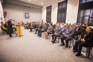 Na dużej sali w kilku rzędach siedzą zaproszeni goście konferencji i słuchają wykładu prelegenta, który stoi po lewej stronie przy mównicy. W pierwszym rzędzie siedzą przedstawiciele służb mundurowych i  władz miasta Gorlic.