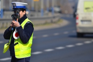 Policjantka ruchu drogowego (widoczna żółta kamizelka, biała czapka) mierzy prędkość ręcznym miernikiem. W tle - ruch uliczny.