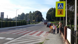 Widoczna droga z przejściem dla pieszych. Na chodniku po jednej stronie stoi policjant, który wręcza odblaski dzieciom idącym do szkoły.