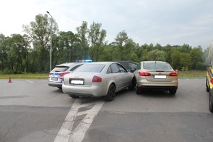 Na skrzyżowaniu drogi stoi samochód który jest zablokowany po uderzeniu pomiędzy innym pojazdem i radiowozem policyjnym.