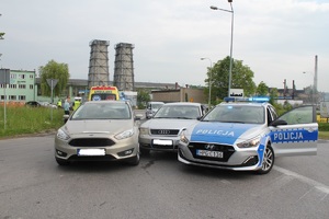 Na skrzyżowaniu drogi stoi samochód który jest zablokowany po uderzeniu pomiędzy innym pojazdem i radiowozem policyjnym. W tle widać ambulans.