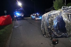 Po prawej stronie na drodze leży na boku samochód z rozbita przednią szybą i pogiętą pokrywą silnika, obok stoi wyjęty akumulator. Nieco dalej na drodze stoi radiowóz policyjny i pojazd straży pożarnej z włączonymi sygnałami świetlnymi uprzywilejowania. Po lewej stronie stoi czerwona kurtyna osłaniająca miejsce wypadku.