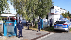 Czterech policjantów stoi przy bramie wjazdowej na teren zakładu. Obok bramy stoi radiowóz.