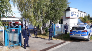 Czterech policjantów stoi przy bramie wjazdowej na teren zakładu. Obok bramy stoi radiowóz.