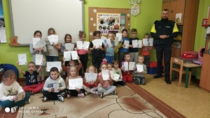 Dzieci stoją w grupie i w rękach trzymają kartki edukacyjne, obok stoi Policjant.