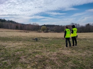 Dwóch policjantów w trakcie uruchamiania drona na tle łąk i lasów.