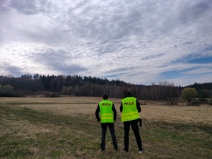 Dwóch policjantów obserwuje z drona tereny narażone na podpalenia. W tle łąki i lasy.