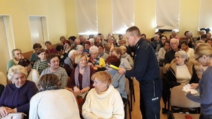 Policjant na dużej sali rozdaje siedzącym seniorom odblaski.