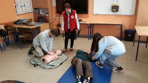 Na podłodze leży osoba, której udzielana jest pierwsza pomoc, obok lezy fantom, na którym młody chlopak wykonuje  sztuczne oddychanie. Całość nadzoruje ratownik medyczny