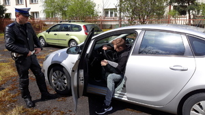 Obok samochodu stoi policjant, w samochodzie są otwarte drzwi kierowcy, na miejscu kierowcy siedzi mlodu mężczyzna.