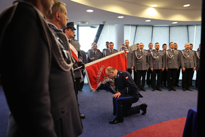 Komendant Powiatowy Policji w Gorlicach wita się ze sztandarem jednostki przyklękując i całując sztanadar. W tle funkcjonariusze Policji.