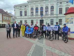 Rowerzyści oraz kilka innych osób stoi trzymając flagi IPA na tle budynku Urzędu Miasta w Sanoku.