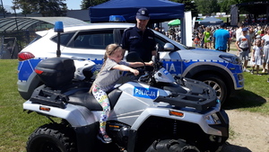 Dziewczynka siedzi na quadzie policyjnym. Obok stoi policjant w tle radiowóz policyjny.