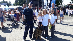 Dzieci głaskają policyjnego psa, który jest razem z przewodnikiem. W tle piknik z okazji dnia dziecka.