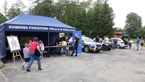 Policyjny stoisko z namiotem z napisem Komenda Powiatowa Policji w Gorlicach. Obok namioty stoją policyjny quad, motocykl i samochód. Do stoiska podchodzą dorośli i dzieci.