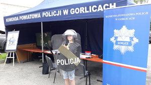 Przy stoisku policyjnym stoi chłopiec ubrany w wyposażenie policjanta, na głowie ma kask, trzyma tarczę i pałkę.