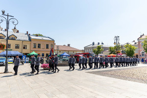 Pododział Policji wchodzi na rynek miasta. W tle stoiska promocyjne służb mundurowych.