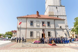 Zaproszeni goście stoją na płycie rynku w Bieczu na tle budynku bieckiego ratusza. Podnoszona jest flaga państwowa.