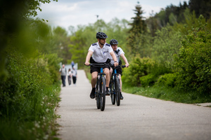 Dwóch policjantów na rowerach patroluje ścieżkę rowerową. W tle idące ścieżką osoby.