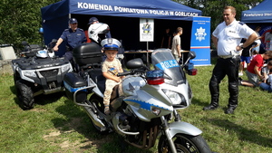 Na motocyklu policyjnym siedzi mały chłopiec w policyjnej czapce. Obok i z tyłu stoją policjanci. W tle policyjny namiot.