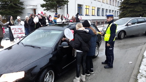 Kilka młodych osób rozmawia z kierowcą pojazdu, obok stoi policjant. W tle stoi grupa młodzieży.