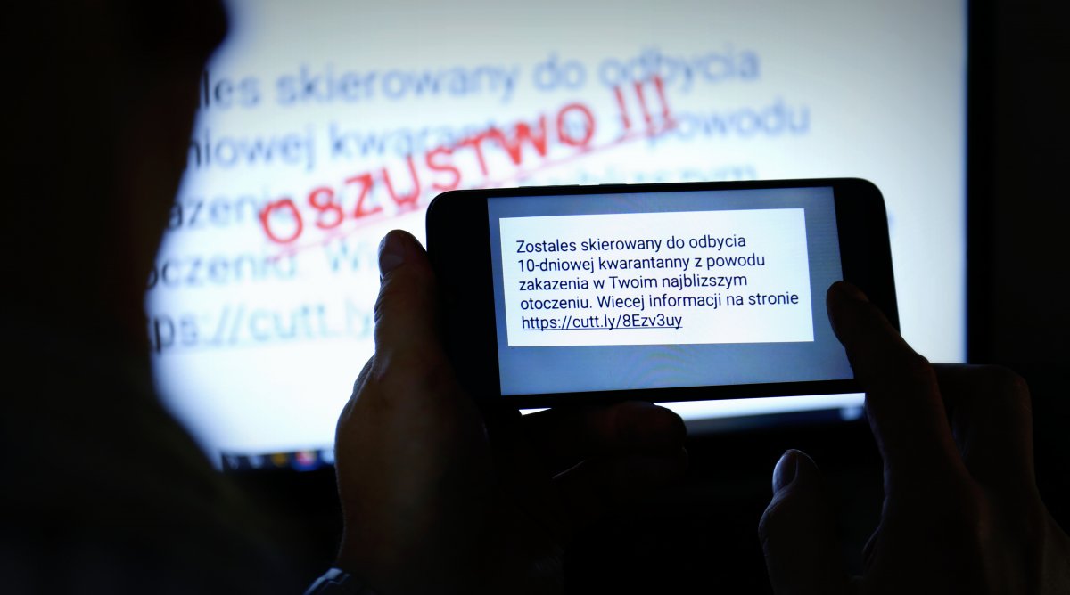 W pierwszym planie zdjęcia widzimy telefon komórkowy trzymany w rękach z wyświetlającym się napisem stworzonym przez oszustów o obowiązku 10 dniowej kwarantanny.